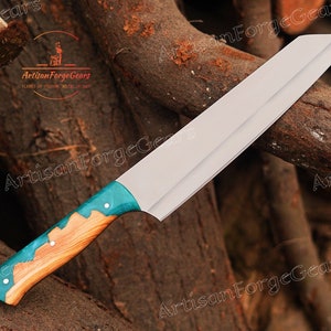 May include: A long, silver chef's knife with a wooden handle. The handle is partially covered in a turquoise blue epoxy resin. The knife is resting on a pile of wood.