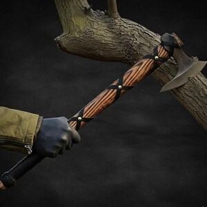 May include: A handcrafted axe with a wooden handle featuring a dark grain pattern. The handle is wrapped with black leather straps secured by brass rivets. The axe head is made of metal with a curved blade. The axe is held against a dark background.