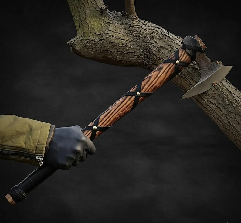May include: A handcrafted axe with a wooden handle featuring a dark grain pattern. The handle is wrapped with black leather straps secured by brass rivets. The axe head is made of metal with a curved blade. The axe is held against a dark background.