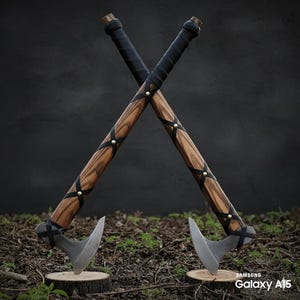 May include: Two crossed axes with wooden handles and metal blades. The handles are wrapped in black leather and adorned with brass studs. The axes are displayed on a bed of grass and small tree stumps.