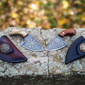 Könnte beinhalten: Zwei kleine, dekorative Messer mit Holzgriffen und gemusterten Klingen, jeweils mit einer Lederscheide. Ein Messer hat ein Schneeflockenmuster, das andere ein dreieckiges Symbol. Die Scheiden sind braun und marineblau, mit Metallakzenten.