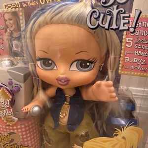 Pode incluir: Uma boneca Bratz Babyz com cabelo loiro e mechas azuis, vestindo uma roupa dourada e azul, segurando um microfone. A boneca est&aacute; em p&eacute; na frente de uma m&aacute;quina de karaok&ecirc; rosa e branca com as palavras "Babyz The Movie Crazy Karaoke" nela. O fundo &eacute; uma caixa rosa e branca com as palavras "So Cute!" e "5 Exclusive Songs from Bratz Babyz the Movie!" nela.