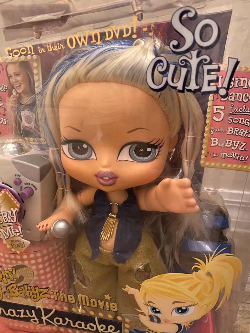 Pode incluir: Uma boneca Bratz Babyz com cabelo loiro e mechas azuis, vestindo uma roupa dourada e azul, segurando um microfone. A boneca est&aacute; em p&eacute; na frente de uma m&aacute;quina de karaok&ecirc; rosa e branca com as palavras "Babyz The Movie Crazy Karaoke" nela. O fundo &eacute; uma caixa rosa e branca com as palavras "So Cute!" e "5 Exclusive Songs from Bratz Babyz the Movie!" nela.