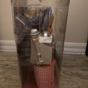 Pode incluir: Uma embalagem de pl&aacute;stico transparente contendo uma boneca com cabelo azul e branco, vestindo um vestido de bolinhas rosa e branco. A boneca est&aacute; segurando um microfone e est&aacute; em p&eacute; em uma plataforma de bolinhas rosa e branco.