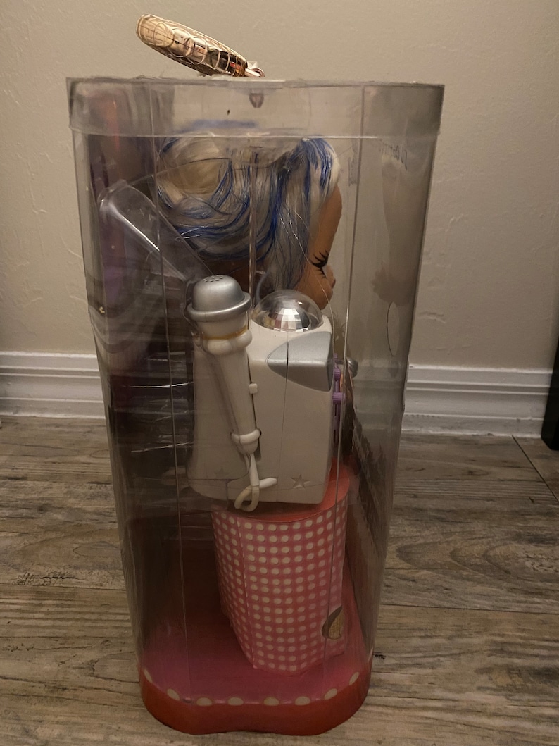 Pode incluir: Uma embalagem de pl&aacute;stico transparente contendo uma boneca com cabelo azul e branco, vestindo um vestido de bolinhas rosa e branco. A boneca est&aacute; segurando um microfone e est&aacute; em p&eacute; em uma plataforma de bolinhas rosa e branco.