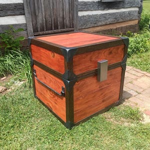 May include: A rectangular wooden chest with a reddish-brown finish, featuring dark brown wooden trim along the edges and corners. The chest includes black metal corner reinforcements, a central silver-toned metal latch, and a black metal side handle.