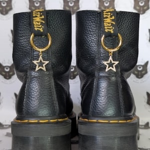 May include: A pair of black leather boots with a platform sole. The boots have yellow stitching and a gold-colored ring with a star charm. The brand name is printed on a black strap.