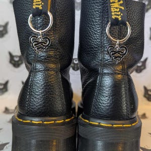 Puede incluir: Botas negras de cuero Doc Martens con costuras amarillas y una suela gruesa de plataforma. Las botas tienen un dije plateado sujeto a los cordones que presenta un corazón con un diseño de nudo. El dije está sujeto a un anillo plateado.