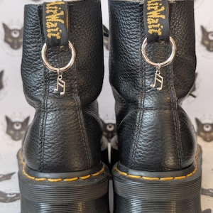 Pode incluir: Um par de botins de couro preto com acabamento texturizado e solas de borracha grossas. Cada bota apresenta uma costura amarela na sola e uma aba preta no calcanhar com texto amarelo. Um anel de metal prateado com um pingente de nota musical.