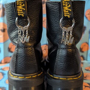 May include: Black leather boots with yellow stitching and thick soles. The boots have a chain with a charm that says "RUM" on one boot and "HAM" on the other. The boots have a yellow tag that says "Dr. Martens" on each boot.