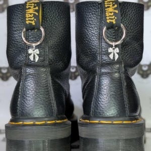 May include: Pair of black leather platform boots with yellow stitching and a pull tab with "Air Wair" text. Each boot has a silver ring and a small bow charm attached. The boots have a textured leather appearance.