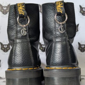 Pode incluir: Um par de botas de cano curto em couro preto com solas de plataforma de borracha grossa e costuras amarelas. Cada bota apresenta uma aba de calcanhar preta com texto AirWair amarelo, um anel de metal prateado e um pingente de letra G.