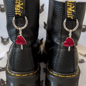 May include: Black leather boots with a yellow stitch detail and a red heart charm attached to a silver chain. The boots have a Dr. Martens logo on the back.