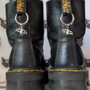Puede incluir: Par de botas de cuero negro con suela de plataforma. Cada bota tiene un dije de murciélago plateado colgando del lazo de la lengüeta. Los lazos son negros con texto amarillo.