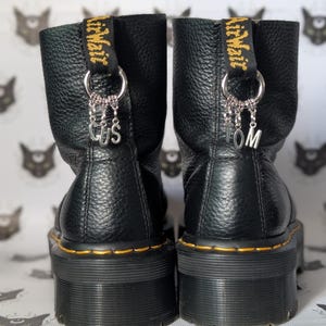 May include: Black leather platform boots with yellow stitching and a black and gold strap with the text "AirWair" on each boot. The boots have silver charms with the letters "CUS" and "TOM" hanging from them.