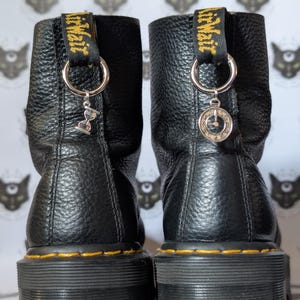 Könnte beinhalten: Ein Paar schwarze Lederstiefeletten mit genarbter Textur und gelben Nähten entlang der dicken Gummisohlen. Jeder Stiefel hat eine schwarze Fersenschlaufe mit gelbem Text und einen silbernen Metallring, an dem ein kleiner Anhänger befestigt ist.