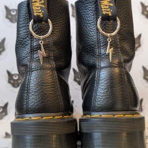 Puede incluir: Un par de botines de cuero negro con acabado texturizado, con suelas gruesas negras y costuras amarillas. Cada bota tiene una trabilla negra en el talón con texto amarillo y una anilla metálica plateada con un dije de rayo plateado.