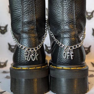 Puede incluir: Botas de cuero negro con suelas gruesas y cadenas plateadas con dijes "F**K" y "OFF" adjuntos.