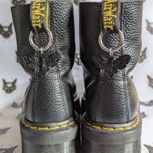 Może przedstawiać: Czarne skórzane buty platformowe z żółtymi szwami i czarnym zawieszką w kształcie ćmy na każdym bucie. Buty mają żółto-czarną metkę "Dr. Martens" z tyłu.