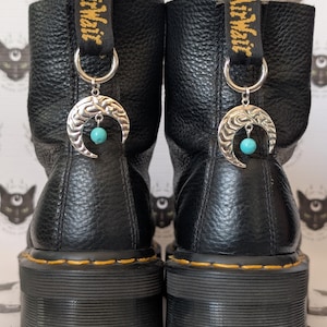 May include: Pair of black leather boots with yellow stitching and platform soles. Each boot features a silver crescent moon charm with a turquoise bead. The charms are attached to the black and yellow pull tabs.