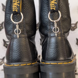 Könnte beinhalten: Ein Paar schwarze Lederstiefeletten mit genarbter Textur und dicken schwarzen Sohlen mit gelben Nähten. Jeder Stiefel hat eine schwarze Fersenzuglasche mit gelbem Text und einen silberfarbenen Metallring-Anhänger in Form eines Pfeils.