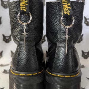 May include: Two black leather platform boots with yellow stitching and a pull tab with the text "AirWair". Each boot features a silver sword charm suspended from a metal ring.