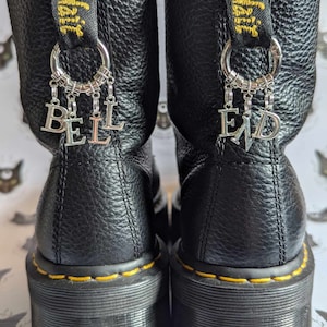 Könnte beinhalten: Schwarze Lederstiefel mit gelben Nähten und dicken Sohlen. Die Stiefel haben silberne Anhänger mit den Worten "BELL END", die daran hängen.