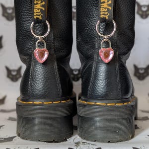 Puede incluir: Un par de botas de plataforma de cuero negro con dijes de cerradura en forma de corazón rosa. Las botas tienen costuras amarillas y un acabado texturizado. Las presillas dicen 'AirWair'. El fondo tiene un patrón de caras de gato.