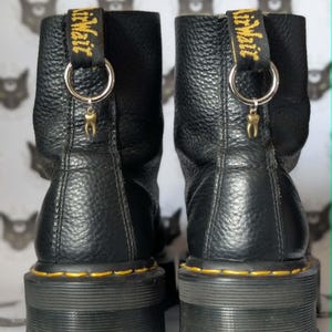 Puede incluir: Par de botines de cuero negro con suelas gruesas y costuras amarillas. Cada bota tiene una lengüeta amarilla y negra con la inscripción "Air Wair" y un pequeño dije de diente dorado.