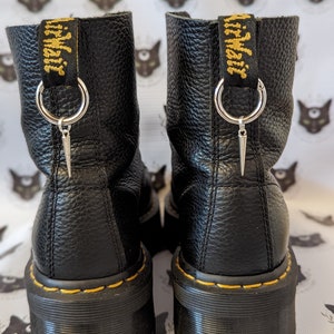 May include: Pair of black leather platform boots with yellow stitching and a pull tab with "Dr. Martens" in yellow text. Each boot has a silver ring with a silver spike charm. The boots have a textured leather finish.