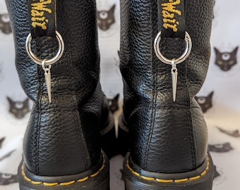 May include: Pair of black leather platform boots with yellow stitching and a pull tab with "Dr. Martens" in yellow text. Each boot has a silver ring with a silver spike charm. The boots have a textured leather finish.