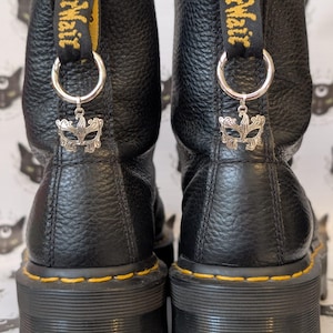 Könnte beinhalten: Ein Paar schwarze Lederstiefel mit strukturierter Oberfläche und gelben Nähten an den dicken Sohlen. Jeder Stiefel hat eine schwarze Fersenschlaufe mit gelbem AirWair-Schriftzug, einen silbernen Metallring und einen silbernen Maskenanhänger.