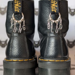 Może przedstawiać: Para czarnych skórzanych butów na platformie z żółtymi przeszyciami i uchwytem z nadrukiem "airwair". Każdy but ma srebrny charms z literami "YUNG" i "BLUD".