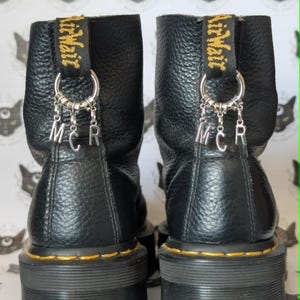 May include: Two black leather ankle boots with chunky, ridged soles. Each boot has a yellow and black pull tab with "Air Wair" and a silver charm with the letters "MCR". The boots have yellow stitching around the soles.