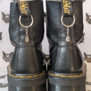 May include: A pair of black leather ankle boots with a textured surface, featuring yellow stitching along the thick rubber soles. Each boot has a black heel pull tab with yellow text and a silver metal ring attached, holding a small guitar charm.