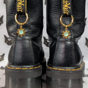 Può includere: Paio di stivali platform in pelle nera con cuciture gialle e linguette. Ogni stivale presenta un cerchio color oro con un ciondolo a forma di sole con un centro azzurro. Gli stivali hanno una tomaia in pelle strutturata e una suola spessa e scanalata.