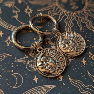 May include: Gold-colored hoop earrings with celestial charms. Each charm features a detailed sun and moon design. The earrings are displayed on a dark background with a celestial pattern, including moons, stars, and suns.