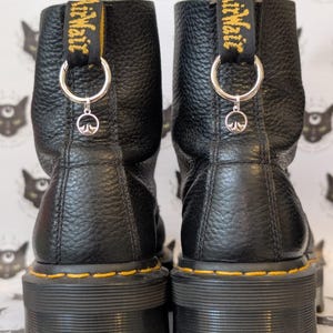 Könnte beinhalten: Ein Paar schwarze Lederstiefel mit strukturierter Oberfläche und dicken schwarzen Sohlen mit gelben Nähten. Jeder Stiefel hat eine schwarze Fersenschlaufe mit gelbem AirWair-Schriftzug, einen silbernen Metallring und einen kleinen silbernen Anhänger.