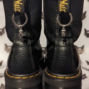 Może przedstawiać: Czarne skórzane buty z żółtymi szwami i srebrnym wisiorkiem przymocowanym do pierścienia na górze każdego buta. Wisiorkiem jest mały ząb.