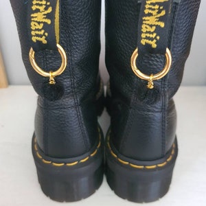 May include: Pair of black leather ankle boots with yellow stitching and gold accents. The boots have a chunky platform sole and a gold charm with a small pom-pom attached.