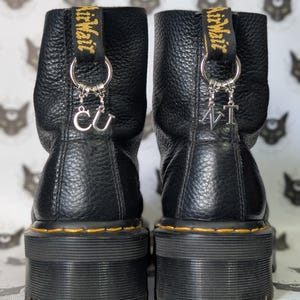 Puede incluir: Botines de cuero negro con suela de plataforma y costuras amarillas. Las botas tienen una lengüeta con texto amarillo y dijes plateados con las letras "CU" y "AT".