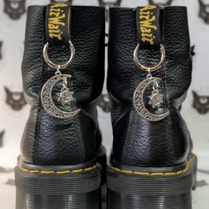 Può includere: Paio di stivaletti in pelle nera con suole platform. Ogni stivaletto presenta un ciondolo argentato con un design a mezzaluna e stella. Gli stivaletti hanno cuciture gialle e un tirante nero e giallo.