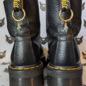 May include: Black leather boots with yellow stitching and a thick platform sole. The boots have a gold ring with a charm attached to the top of each boot. The charm is a small, blue and gold bird.