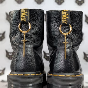 Könnte beinhalten: Schwarze Leder-Ankle Boots mit goldfarbenen Kreuzanhängern. Die Stiefel haben eine klobige Plateausohle mit gelben Nähten und eine schwarze Zuglasche mit goldenem Schriftzug.