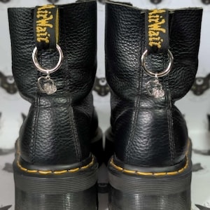 May include: Two black leather ankle boots with yellow stitching and a chunky sole. Each boot features a yellow and black pull tab with "AirWair" text and a silver charm. The boots are set against a patterned backdrop.