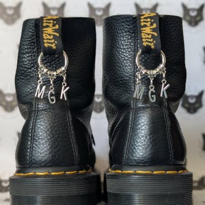 Puede incluir: Botas de plataforma de cuero negro con costuras amarillas y suela negra. Cada bota tiene una lengüeta amarilla y negra con la inscripción "Air Wair". Cuelgan dijes plateados con las letras "MGK".