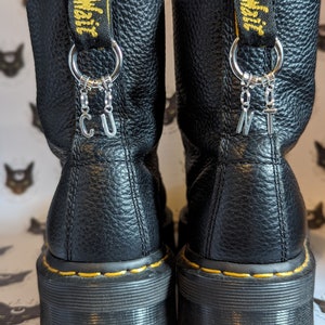 Może przedstawiać: Czarne skórzane buty z żółtymi szwami i grubą platformą. Buty mają srebrny charm z literami "CUNT" wiszącymi z nich.