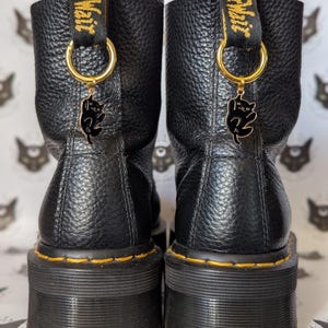 Puede incluir: Par de botas de plataforma de cuero negro con costuras amarillas y presillas. Cada bota tiene un anillo dorado con un dije de gato negro. Las botas tienen un aspecto de cuero texturizado y una suela gruesa y acanalada.