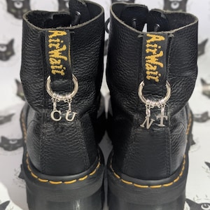 Könnte beinhalten: Schwarze Lederstiefel mit gelben Nähten und den Worten "Air Wair" auf den Zuglaschen. Silberne Anhänger mit den Buchstaben "CU" und "NT" hängen an den Stiefeln.