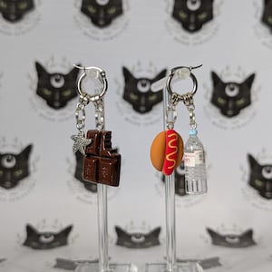 May include: A pair of silver hoop earrings with charms. One earring features a chocolate bar and a starfish charm. The other earring has a hot dog charm and a miniature water bottle charm. The background has a repeating pattern of black cat faces.